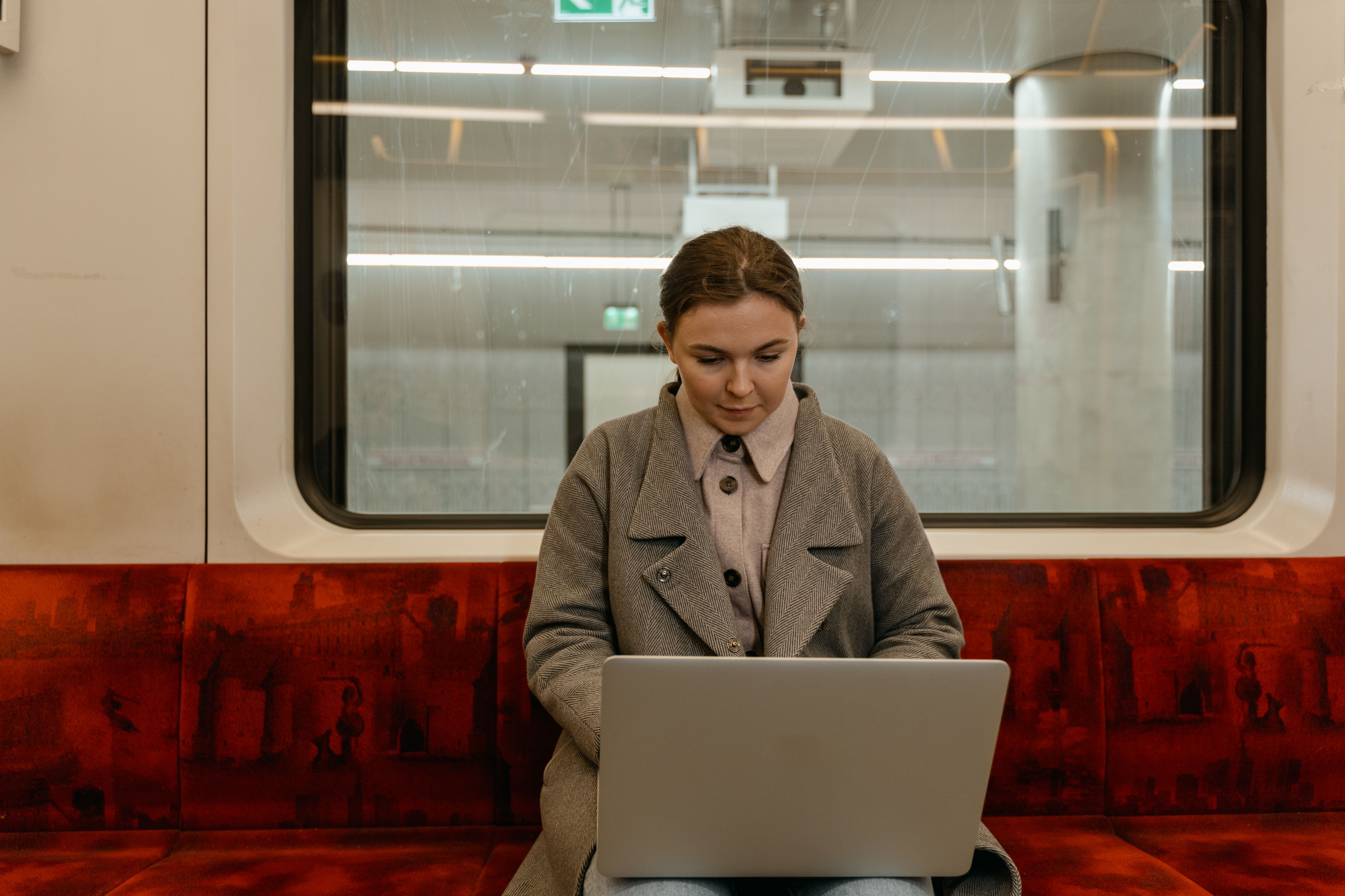 Caucasian woman using laptop in a gray coat sitting inside a subway train.