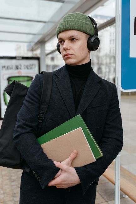 Man in beanie with headphones, holding books at an outdoor transit station. Man in beanie with headphones, holding books at an outdoor transit station.