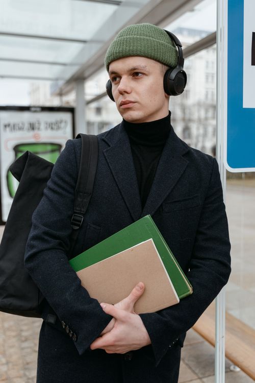 Free Man in beanie with headphones, holding books at an outdoor transit station. Stock Photo
