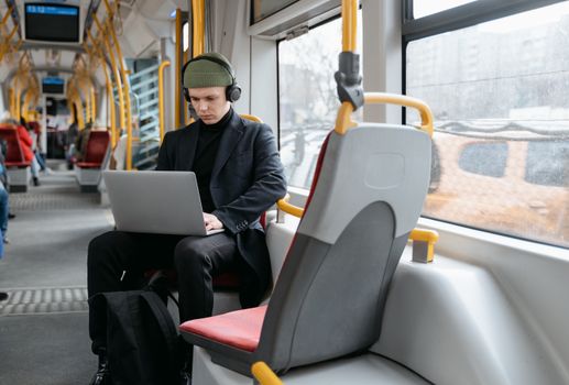A man in a black coat using a laptop while commuting on a tram, listening to music.