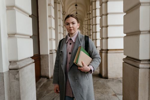 Free A young woman in a gray coat holding books, standing in an elegant academic hallway. Stock Photo
