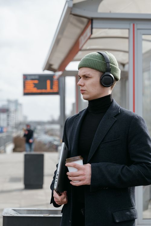 Free A man in a beanie and headphones holding coffee and a laptop outdoors. Stock Photo