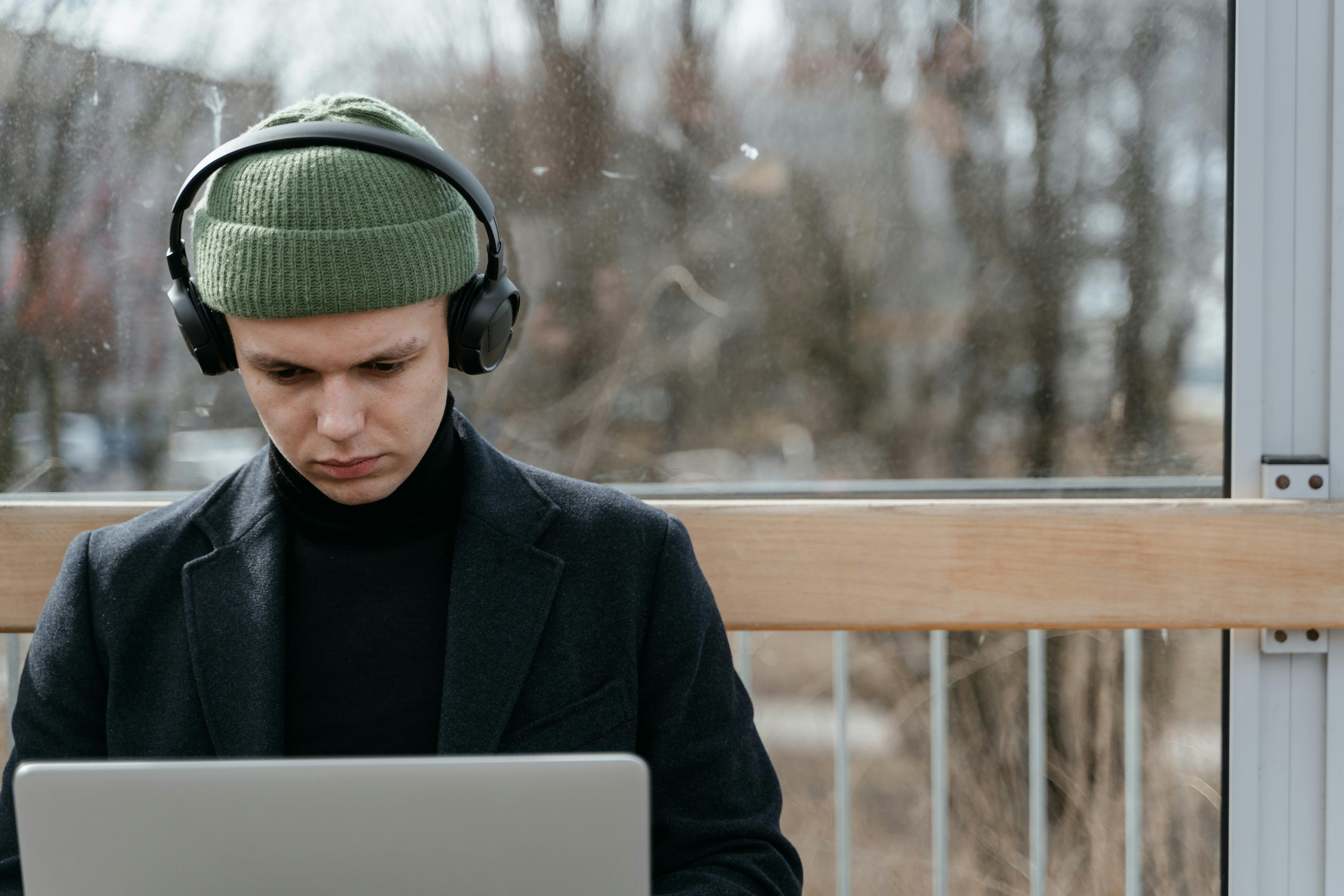 Man wearing green beanie and headphones works on laptop in a bright outdoor setting.