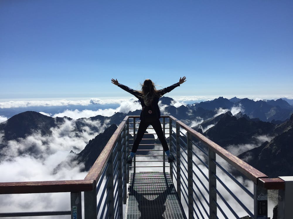 Persona De Pie Sobre Pasamanos Con Los Brazos Abiertos Mirando Hacia Las Montañas Y Las Nubes