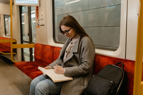 Free A woman in glasses and a gray coat writes in a notebook while commuting on a train. Stock Photo