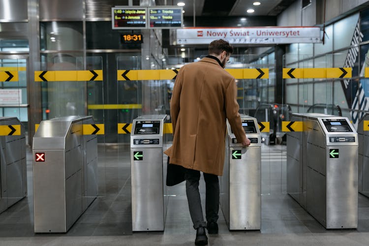 A Back View Of A Man In Brown Coat Standing Between Ticket Barriers