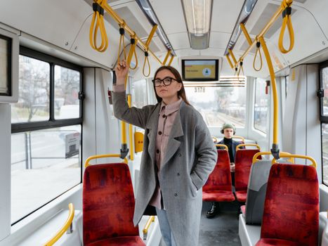 A woman in a coat and eyewear standing inside a public tram, holding onto the handrail.