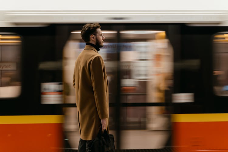 A Man In Brown Coat Standing On A Train Station Near A Moving Train