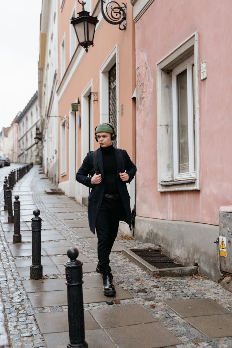 A Man In Black Coat With Headphones Walking On The Sidewalk