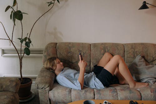 A Boy in Blue Shirt Using a Mobile Phone while Lying on the Sofa