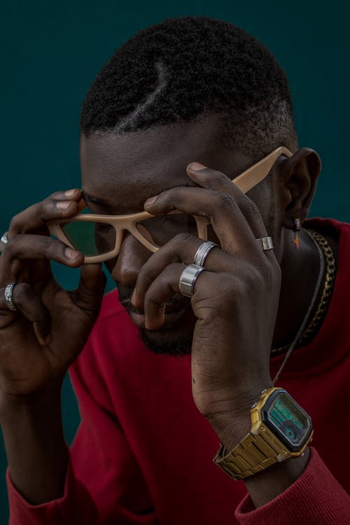 Trendy African American male with hands on modern eyewear wearing wristwatch and rings while sitting in modern studio near wall