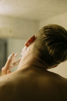 A young man applies shaving cream on his face in a bathroom mirror.