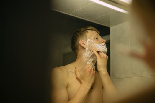 Adult man applying shaving cream while looking in bathroom mirror.