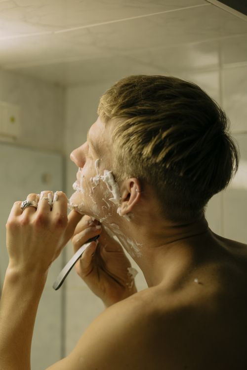 Free Shirtless man with blond hair shaving using a straight razor, focusing on self-care. Stock Photo