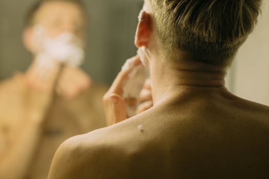 Back view of a man shaving in front of a bathroom mirror, focusing on reflection.