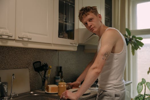 A tattooed man in a white tank top making breakfast in a home kitchen.