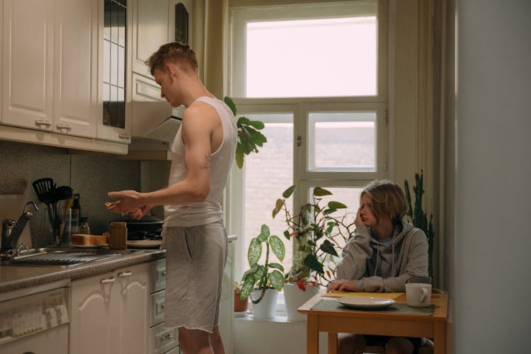 A Young Boy In Gray Sweater Sitting Near The Table While Looking At His Brother Standing Near The Kitchen Cabinets