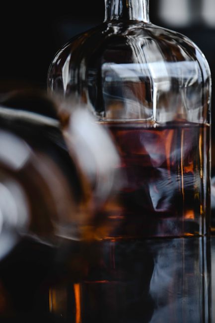 A snifter of amber cognac next to an open bottle, with an oak aging barrel visible in the background.
