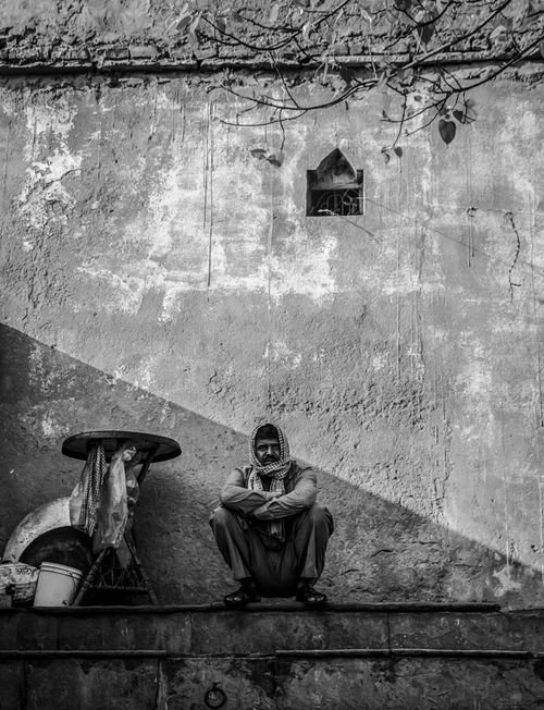 Free A monochrome image of a man crouching against a textured wall in Old Delhi, capturing street life. Stock Photo