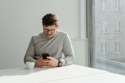 Free Man in gray sweater checking smartphone indoors by a window with a ceramic cup on the table. Stock Photo