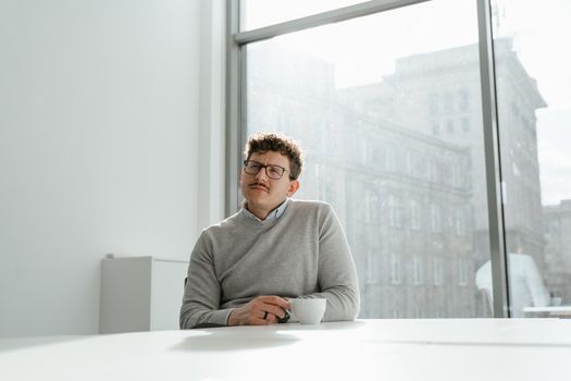 Man in gray sweater thinking with coffee in hand by large window indoors.