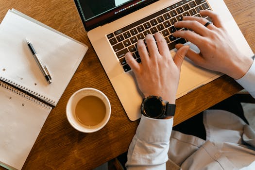 Hands typing on a laptop next to a cup of coffee and a notebook on a wooden table.