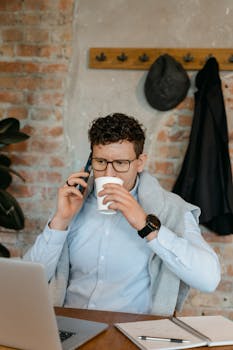 A focused man working remotely, engaged in a phone call while sipping coffee in a casual workspace.