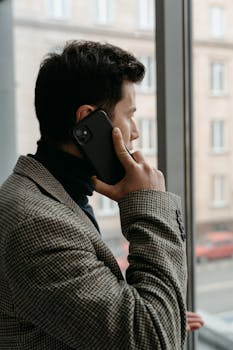Man in a checkered blazer using a smartphone while gazing out a window.