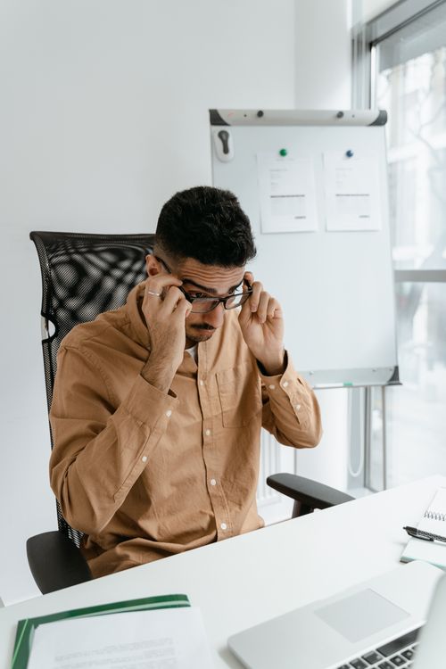 Free A man adjusts his glasses while seated at an office desk beside a window. Stock Photo