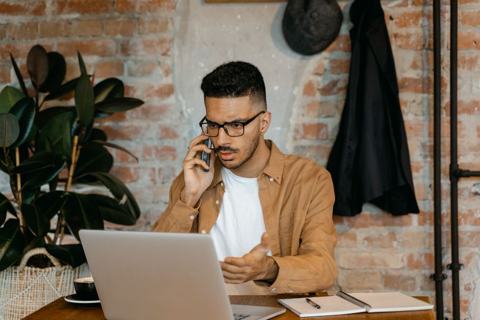 A man in eyeglasses works on a laptop while talking on the phone in a stylish brick-walled office. A man in eyeglasses works on a laptop while talking on the phone in a stylish brick-walled office.