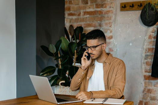 Young man in glasses working on a laptop and phone in a modern office setting.
