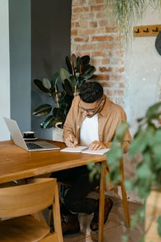 A young man concentrating at a home workspace, taking notes beside a laptop.