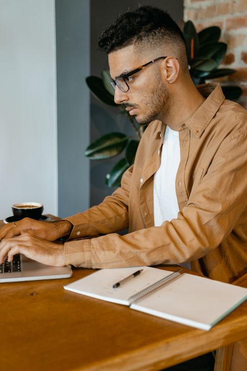 Free Young man with eyeglasses typing on a laptop in a modern cafe setting, perfect for business and freelance themes. Stock Photo