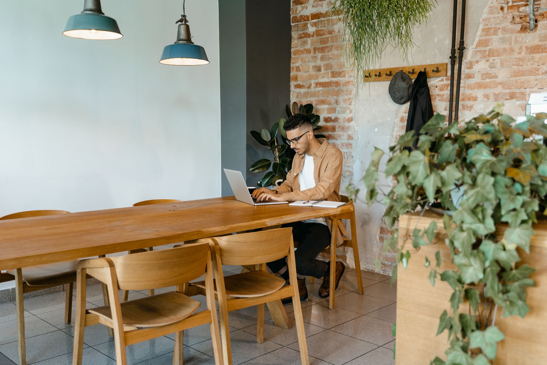 Man typing on laptop in modern, stylish workspace with natural decor and brick wall.