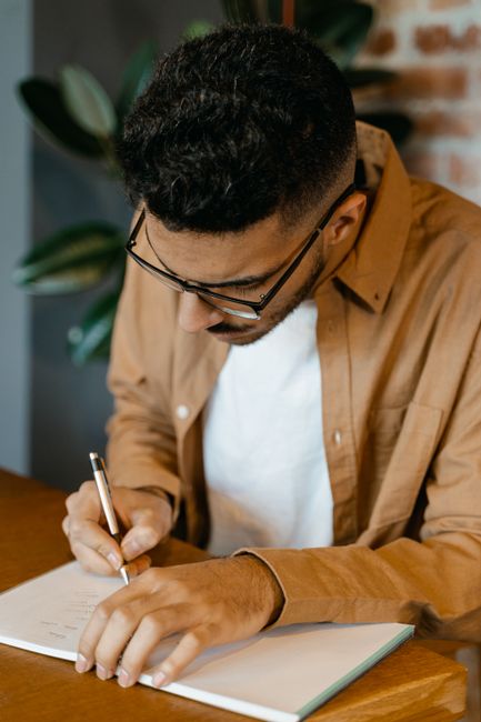 A man in glasses writing in a notebook at a desk, focused and thoughtful. A man in glasses writing in a notebook at a desk, focused and thoughtful.