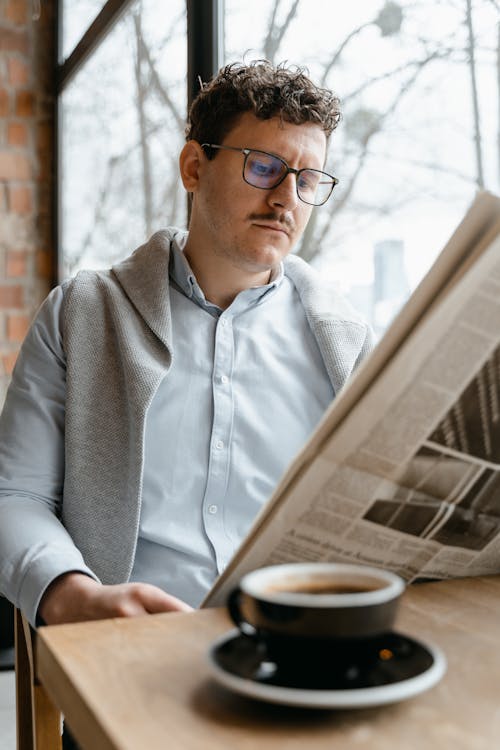 Free Concentrated male in eyeglasses wearing shirt reading newspaper while sitting near window at wooden table with cup of hot coffee Stock Photo