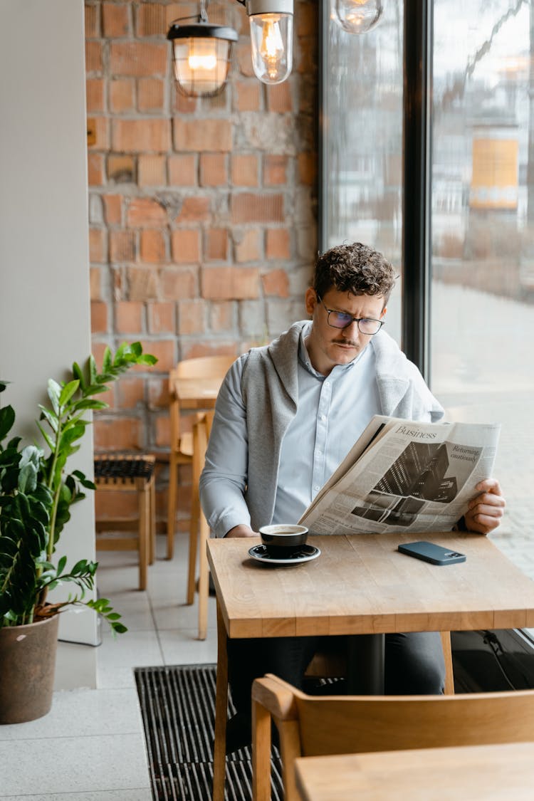 A Man Sitting Beside A Glass Wall Reading Newspaper