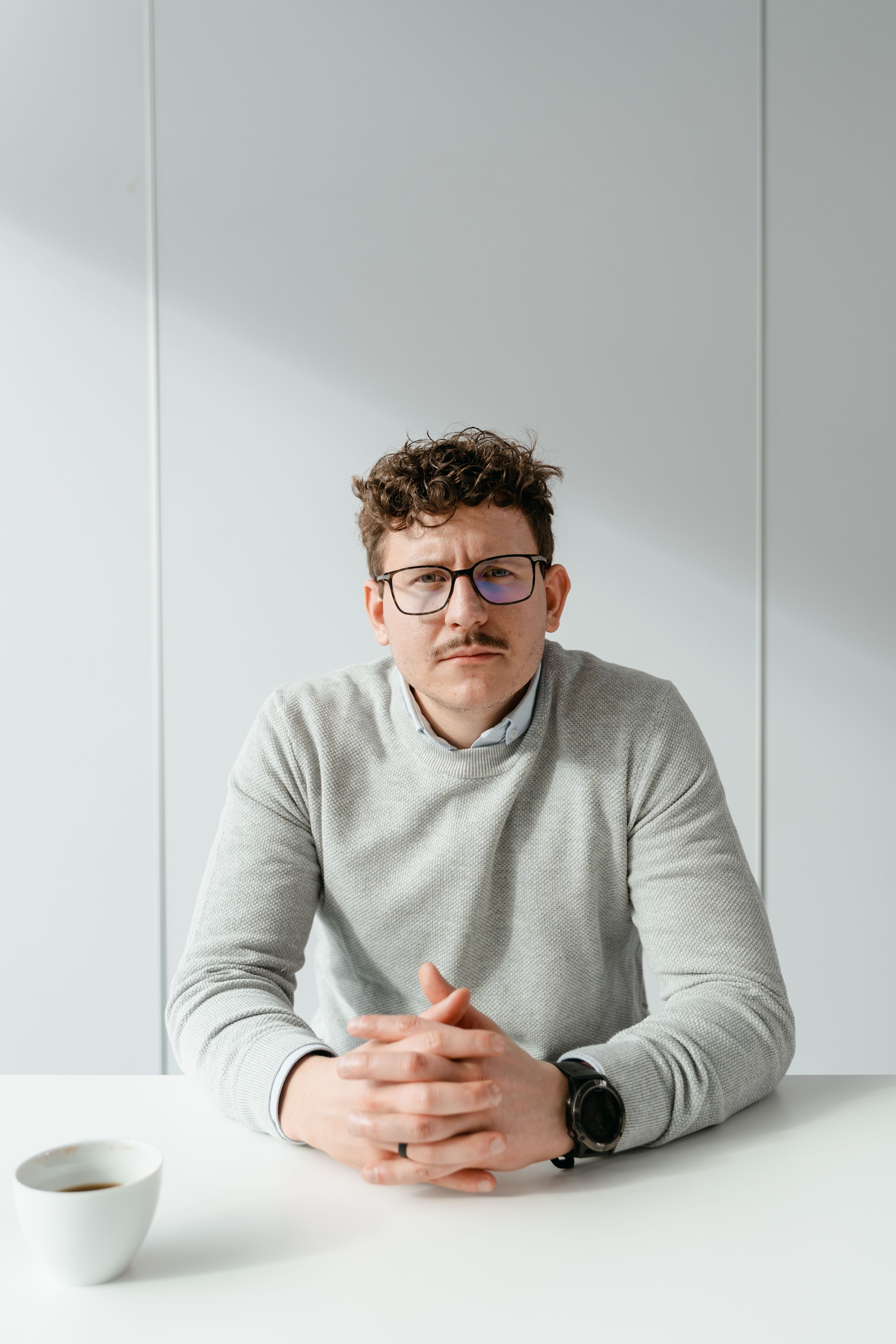 Man with glasses and mustache sitting at a desk with coffee, looking serious.