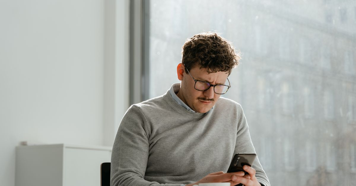 Adult man wearing glasses using smartphone in sunny office space, holding a mug.