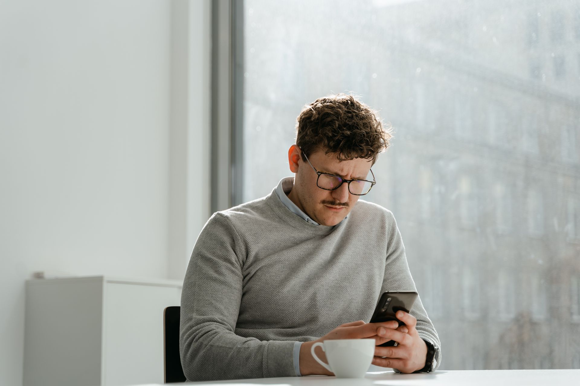 Adult man wearing glasses using smartphone in sunny office space, holding a mug.