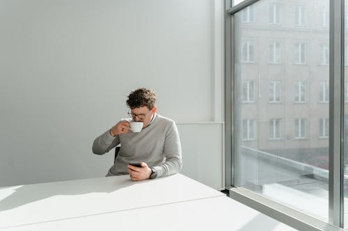 A Man in Gray Sweater Sitting Near the Table while Drinking Coffee