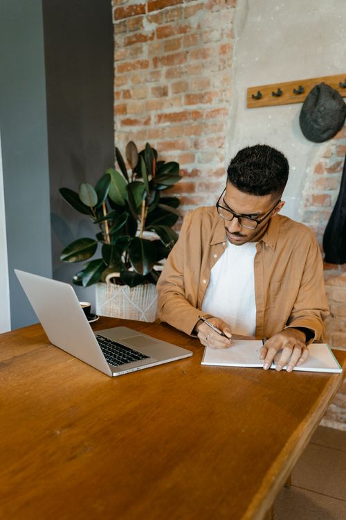 Free Man working in home office writing notes at desk with laptop, indoor plant in view. Stock Photo