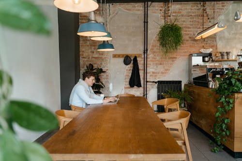 A Man in White Long Sleeves Sitting on a Wooden Chair while Typing on His Laptop