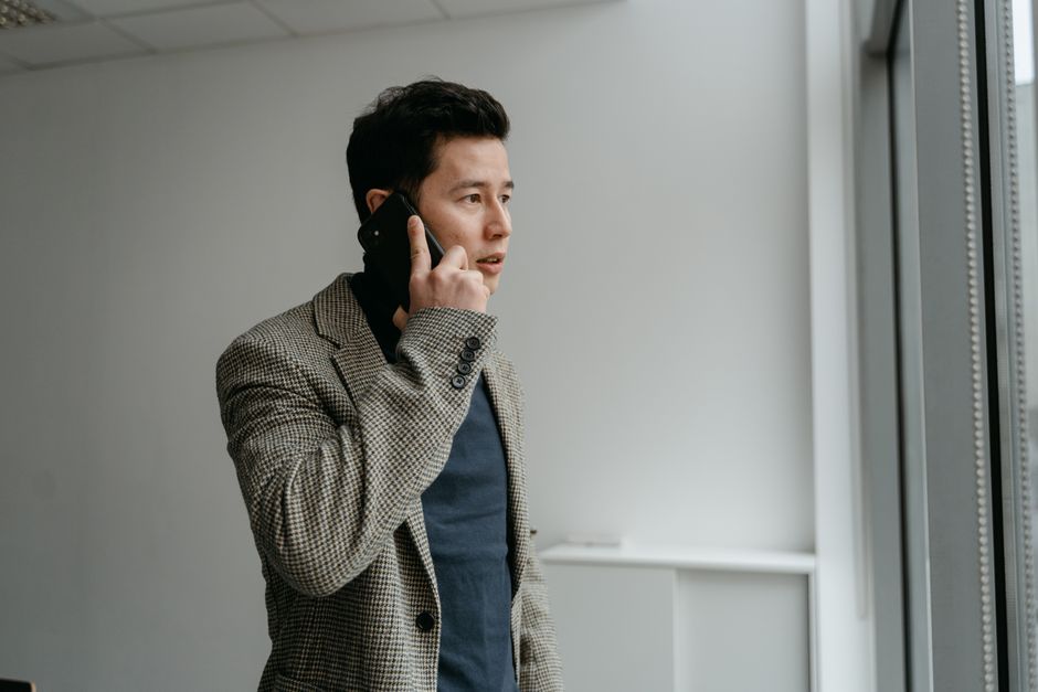 Young businessman in blazer talking on smartphone near window indoors.