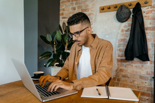 Man in casual office setting typing on laptop with coffee and notebook on desk.
