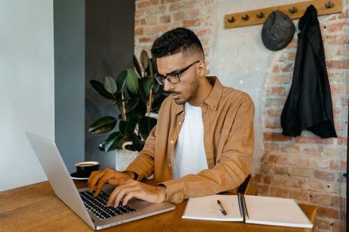 Free Man in casual office setting typing on laptop with coffee and notebook on desk. Stock Photo
