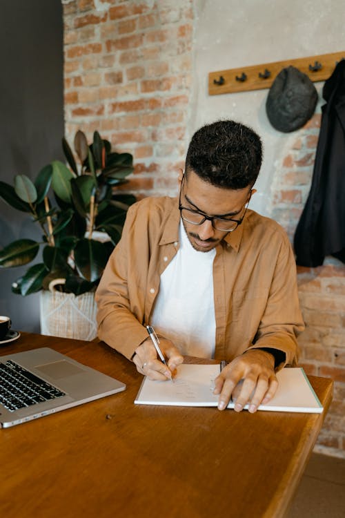 A Man in Brown Long Sleeves Sitting Near the Wooden Table while Writing on His Notebook