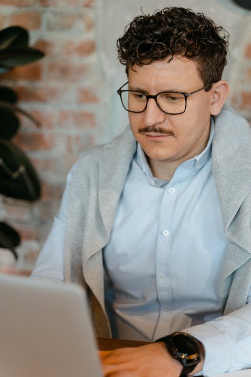 Free A Man in White Long Sleeves Wearing Eyeglasses while Looking at His Laptop Stock Photo