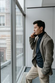 Confident businessman in a gray blazer talking on phone by office window.