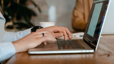 A person typing on a laptop at a wooden table in a modern office setting.